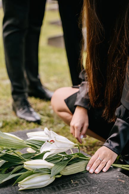 A person placing a white lily bouquet with long green leaves on a dark stone memorial at a funeral site, with other flower arrangements visible in the background. The bouquet features fresh, unopened