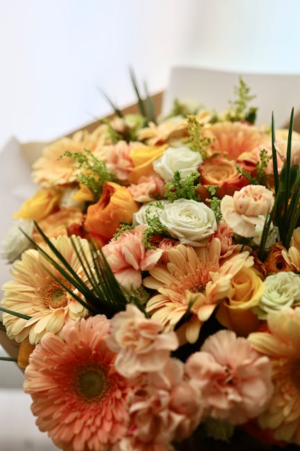 A vibrant floral arrangement featuring a variety of fresh flowers including soft pink gerbera daisies, pale peach roses, and white lisianthus, accented with green foliage and ferns. The bouquet is arr