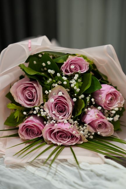 A bouquet of fresh, light pink roses with tightly layered petals, accented by small white baby's breath flowers and surrounded by lush green leaves. The arrangement is carefully wrapped in white trans